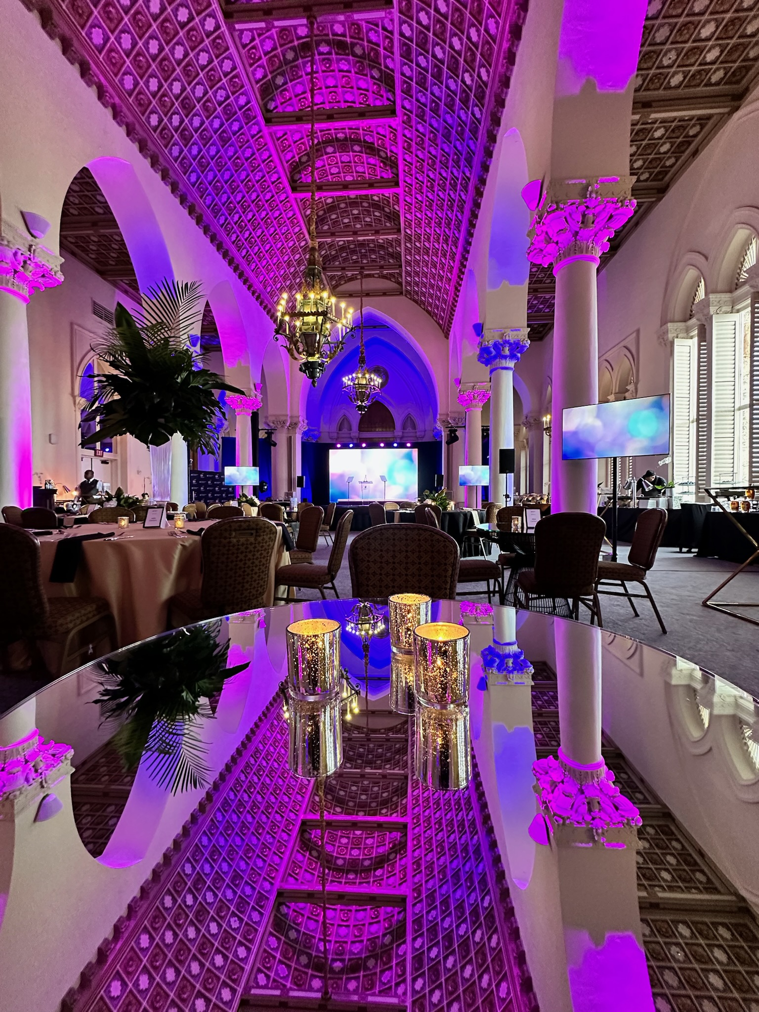 Ornate ballroom with magenta uplighting, chandeliers, and stage screens set for a gala event