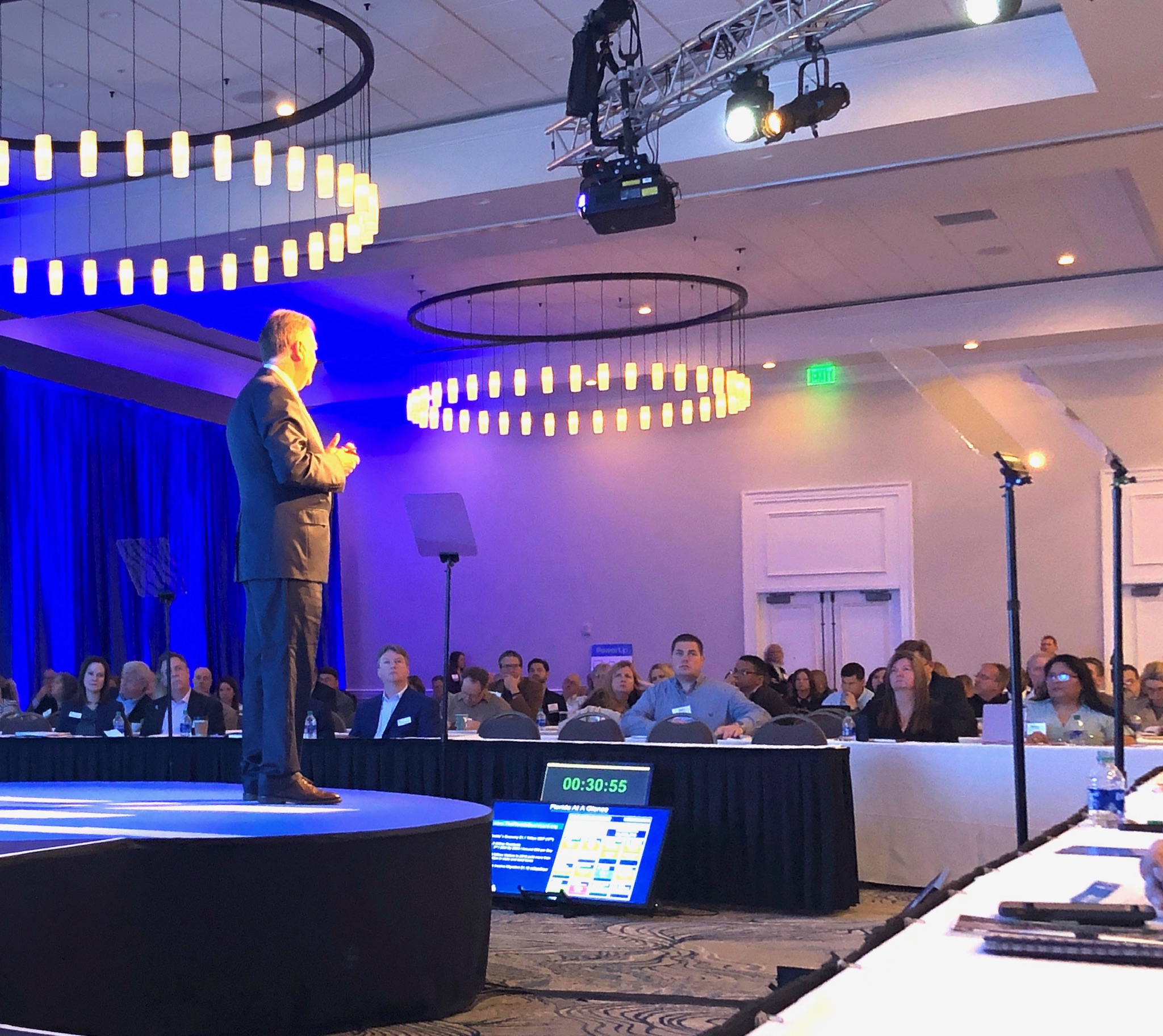 Keynote speaker on a round stage addressing a seated conference audience with a confidence monitor in view